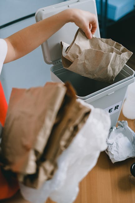 A person wearing a white sleeve is seen placing a crumpled brown paper bag into a white rubbish bin with a black interior, situated on a wooden surface. The bin has a lid that is open, showing a glimp